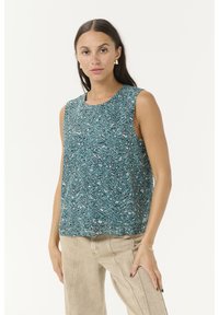 Frau mit langen dunklen Haaren, die ein ärmelloses, t ürkis gemustertes Oberteil, beige Hosen, goldene Ohrringe und ein Armband trägt, steht vor weißem Hintergrund.