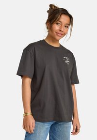 Ung kvinne med håret oppsatt iført grå Billabong Surf Club t-skjorte og blå jeans, smilende litt mot en enkel hvit bakgrunn.
