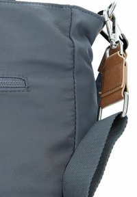 Bolso mensajero de tela gris que cuenta con un bolsillo superior con cremallera, una correa texturizada y un acento de cuero marrón cerca de un cierre de metal.