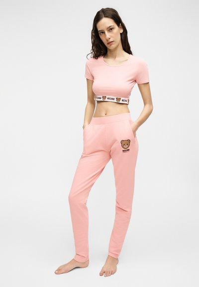 Γυναίκα όρθια ξυπόλητη, που φορά ροζ κοντό τοπ και παντελόνια με λογότυπα αρκούδου στη μέση και στο μηρό, τα χέρια στις τσέπες.