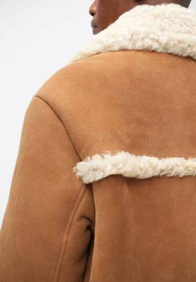 Giacca di camoscio marrone con un colletto peloso crema, caratterizzata da linee pulite e un design strutturato. La texture è liscia con accenti morbidi.