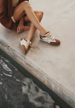 Persona con zapatillas blancas y marrones sentada en un borde con las piernas extendidas sobre el agua, con las manos apoyadas sobre una superficie de hormigón.