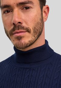 Granatowy sweter dzianinowy z wysokim kołnierzem, z wzorem w warkocze. Tekstura jest prążkowana, a materiał wydaje się miękki.