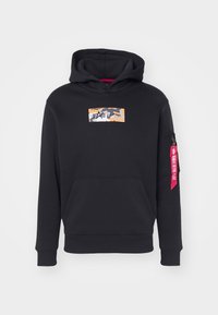 Crna hoodica s kangaroo džepom, narandžasti kamuflažni logo na prsima i džep na rukavu sa patentnim zatvaračem te crvenom oznakom "Remove Before Flight".