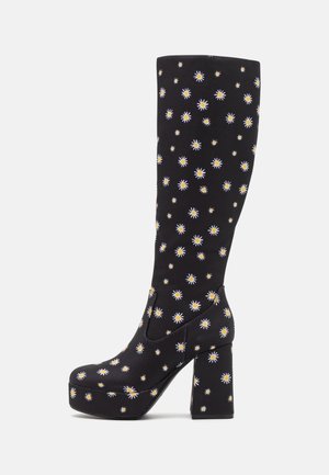 Bottes noires montantes jusqu'aux genoux avec un motif floral présentant des marguerites jaunes et blanches. Fabriquées en matériau souple avec un talon bloc épais et une plateforme.