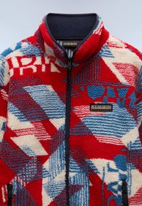 Chaqueta de forro polar con patrón en rojo, azul y crema, con un alto cuello, cierre de cremallera al frente y un parche de logo de tela en el pecho. Superficie texturizada.