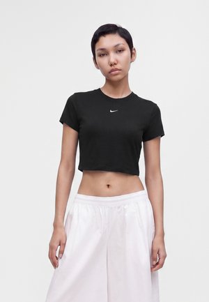Jeune personne aux cheveux courts noirs portant un t-shirt Nike noir court et un pantalon blanc ample, debout devant un fond clair uni.