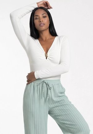 Kobieta z długimi ciemnymi włosami, ubrana w biały top z długim rękawem z detalem na środku oraz jasnoniebieskie spodnie w cienkie paski ze sznurkiem.