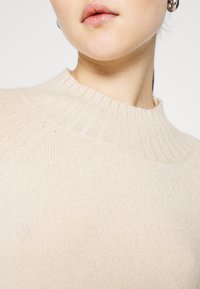 Beige pletený svetr s vroubkovaným kulatým výstřihem a měkkou texturou; detailní pohled na horní část, který ukazuje vymezené švy.