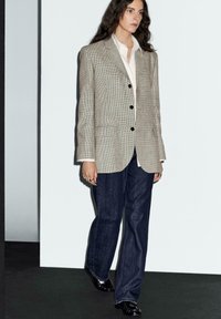 Blazer a quadri beige con bottoni scuri sopra una camicia bianca, abbinato a pantaloni in denim scuri e scarpe nere lucide. Vista a figura intera dell'outfit.