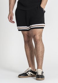 Bermuda nere in maglia con bande orizzontali bianche e marroni all'orlo, abbinate a sneakers nere e beige, caratterizzate da un tessuto testurizzato.