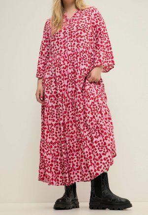 Kobieta ubrana w długą, luźną różową sukienkę w czerwone abstrakcyjne plamy oraz czarne masywne buty, stojąca na tle jednolitego jasnego tła.