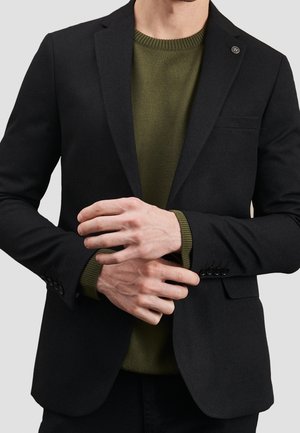 Mand iført en sort blazer over en olivengrøn sweater, der justerer venstre ærme med sin højre hånd mod en ensfarvet baggrund.