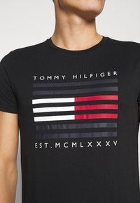 Černé bavlněné tričko s horizontálními pruhy v červené, bílé a námořnické modři, s textem "TOMMY HILFIGER" nahoře a "EST. MCMLXXXV" níže.