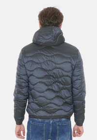 Giacca a piumino grigio scuro con cappuccio, caratterizzata da una texture trapuntata a onde e polsini elastici. Indossata sopra jeans blu, che mostra un look casual.