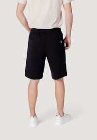 Black cotton shorts with an elastic waistband, two back pockets, and a small logo detail. Worn with a light-colored t-shirt and white sneakers.