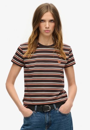 Mujer joven con el pelo largo que lleva una camiseta de rayas en negro, rosa y blanco y vaqueros azules con las manos en los bolsillos, mirando hacia adelante.