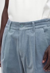 Gros plan d'une personne portant un pantalon en velours côtelé bleu clair avec une ceinture boutonnée et un haut blanc, la main reposant à côté.