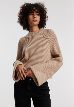 Beige ribgebreide trui met een ronde halslijn, ruime mouwen en een losse pasvorm, gecombineerd met zwarte high-waisted jeans.