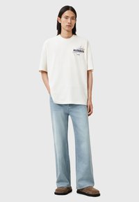 Camiseta oversized beis con un diseño gráfico en el pecho izquierdo, combinada con unos jeans azul claro y calzado marrón tipo slip-on.