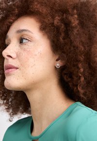 Orecchino a perno in argento con un design circolare e un bordo texturizzato, indossato da una persona con capelli ricci color castano ramato, vestita con una maglietta verde.