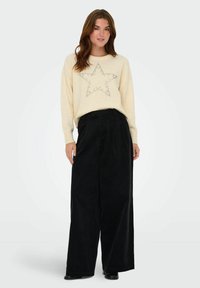 Crème sweater met een sterpatroon van rhinestones, gecombineerd met zwarte, wijde corduroy broeken. Soepele texturen met een relaxte pasvorm.