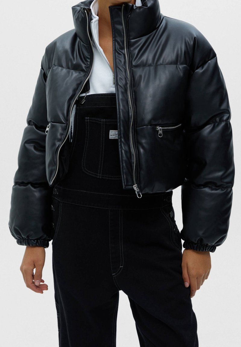Personne portant une veste courte en cuir noir matelassé sur une salopette en denim noir avec des coutures blanches et un haut blanc en dessous.