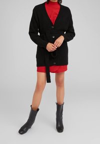 Persona vistiendo un cárdigan negro con cinturón sobre un vestido a cuadros rojos y botas negras hasta el tobillo, de pie contra un fondo gris sencillo.