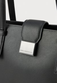 Melnas ādas rokas soma ar teksturētu virsmu, taisnstūrveida sudraba apdari un gludu ādas siksnu.