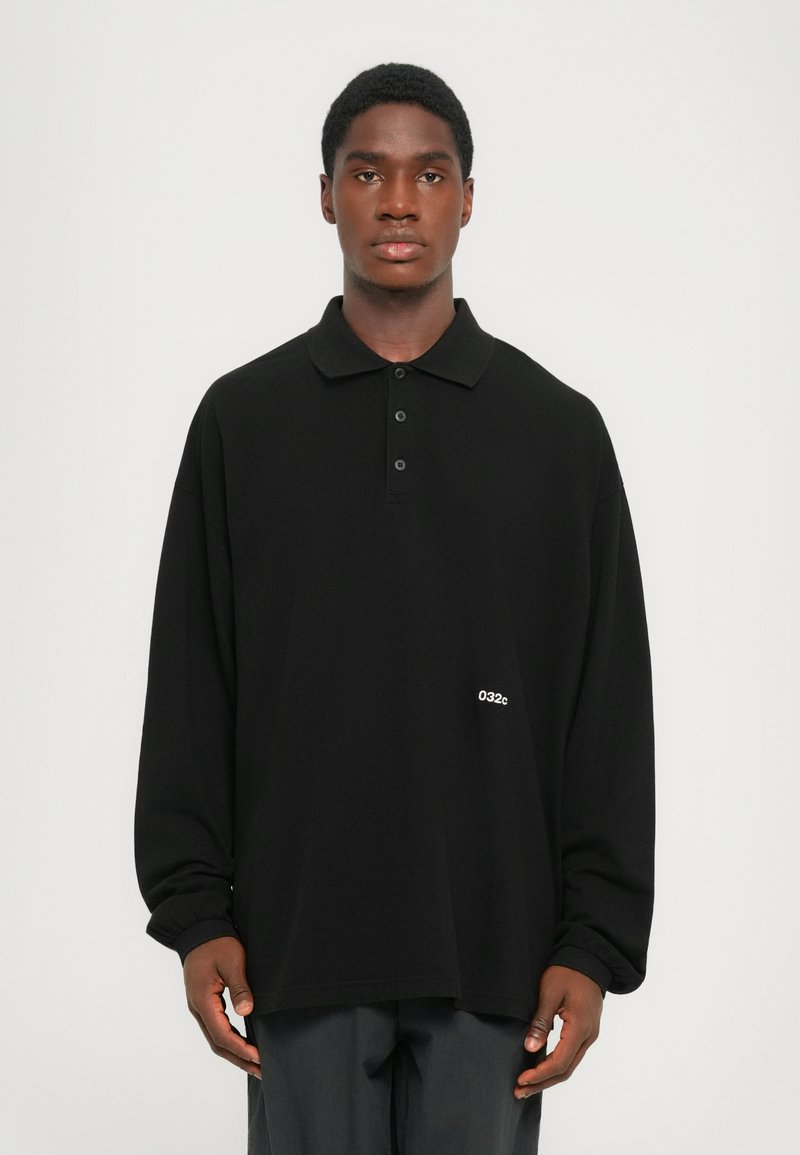 Polo shirt nero a maniche lunghe con tre bottoni e vestibilità ampia. Testo bianco "032c" situato a sinistra vicino all'orlo. Il tessuto sembra morbido e liscio.