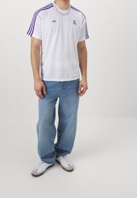 Vit sporttröja med lila ränder och logotyper, kombinerad med ljusblå avslappnade jeans och vita sneakers med grå accenter.