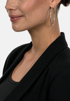 Silver hoop earring with a textured surface, paired with a black blazer and top, highlighting its circular shape and reflective finish.