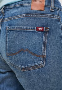 Dżinsy w średnim niebieskim odcieniu z kontrastującymi pomarańczowymi szwami, z tylną kieszenią z czerwoną metką z białym logo konia.