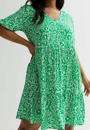 Donna che indossa un vestito verde con motivo floreale bianco, maniche corte, scollo a V e vita arricciata, in piedi con una mano sui fianchi.