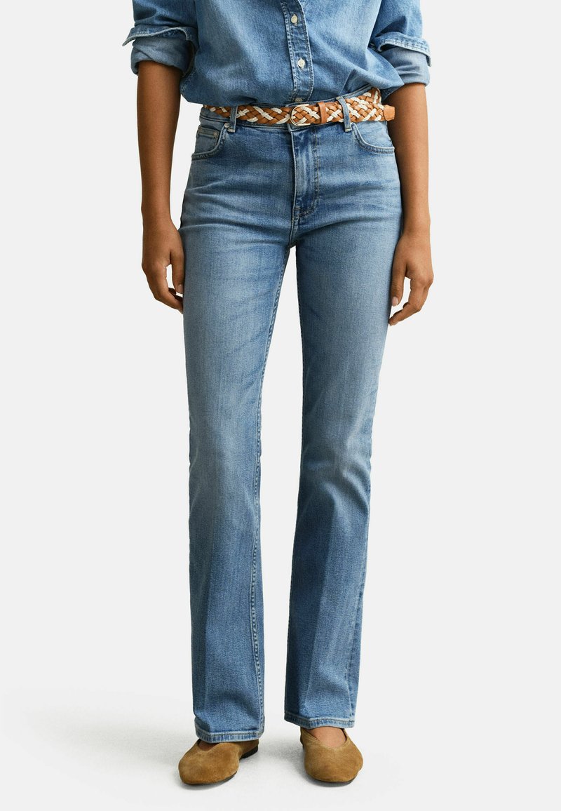 Tenue décontractée avec un jean en denim bleu clair, une ceinture tressée marron, une chemise en denim assortie aux manches retroussées, et des chaussures marron clair sans lacets.
