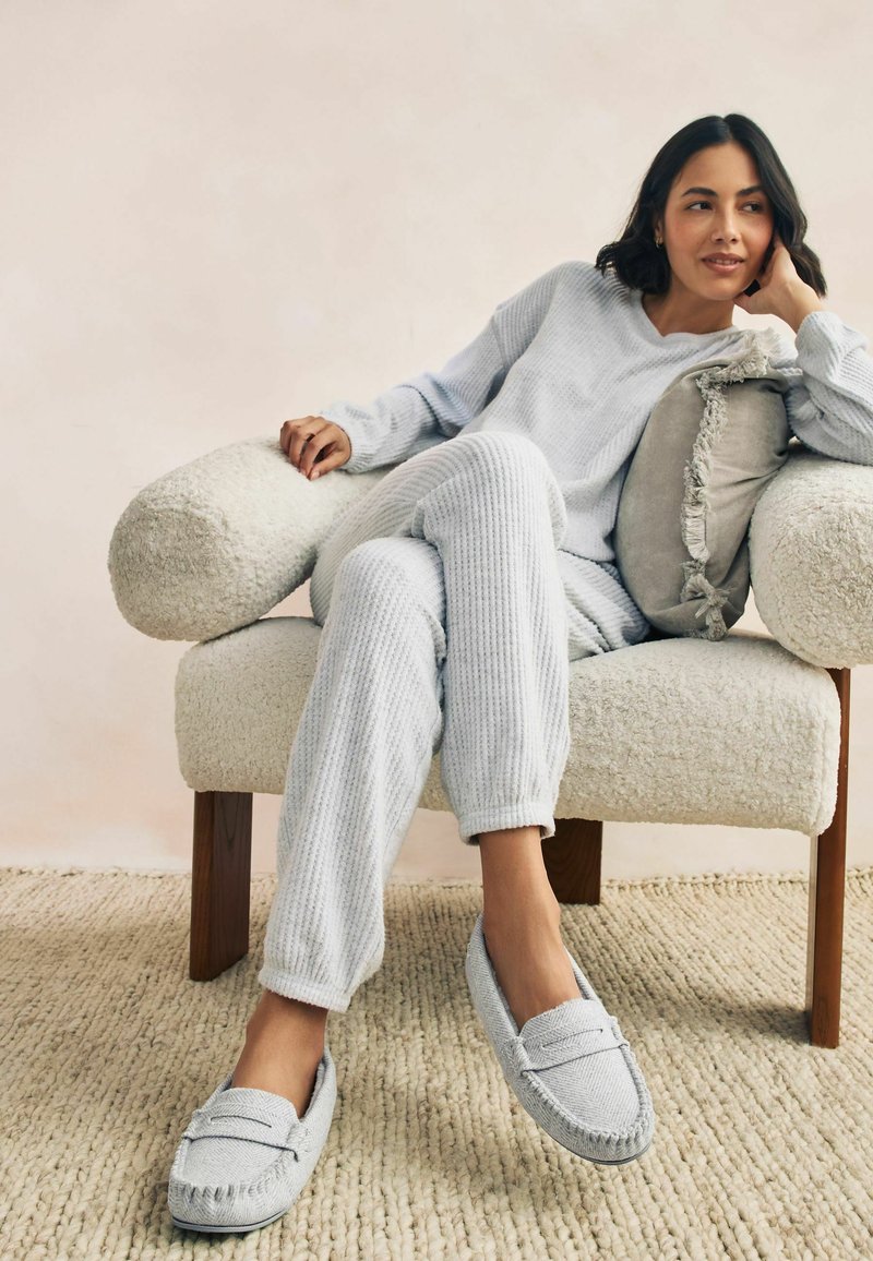 Una donna in abbigliamento comodo grigio chiaro e pantofole abbinate è seduta rilassata su una poltrona texturizzata con un cuscino grigio con frange.
