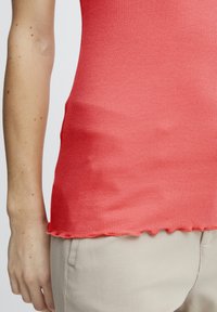 Top in coral aderente e a costine con orlo scallop, realizzato in tessuto elasticizzato, abbinato a pantaloni beige chiaro.