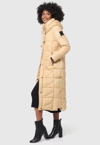 Long beige puffer coat with a hood, featuring quilted patterns, drawstring accents, side pockets, and a slightly fitted silhouette.