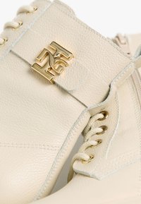 Les bottines en cuir beige présentent une surface texturée, des détails en métal doré, et un design à lacets avec des accents de couture prononcés.