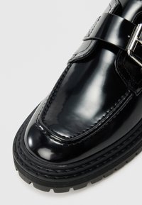 Pantof din piele lac neagră, cu vârf rotund, prevăzut cu o curea lată și o cataramă metalică. Talpă de cauciuc cu profil texturat pentru aderență.