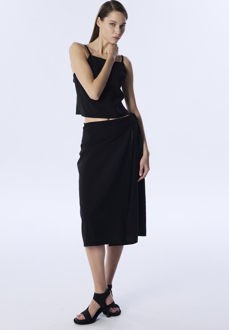 Un crop top noir avec des bretelles fines assorti à une jupe portefeuille arrivant au genou. La tenue présente une texture lisse et un design minimaliste. Des sandales à lanières noires complètent le look.