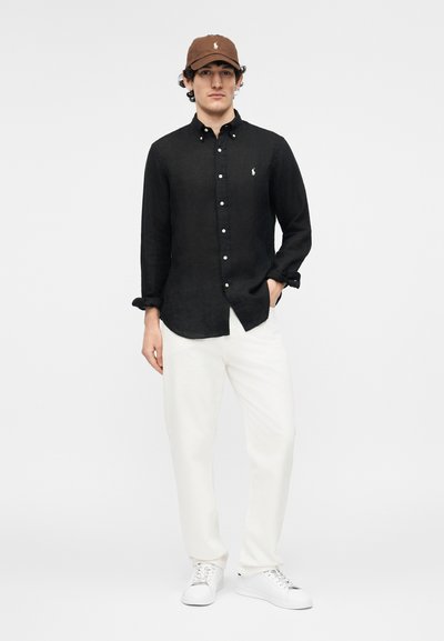 Hombre con gorra marrón, camisa negra de manga larga con botones, pantalones blancos y zapatillas blancas de pie frente a un fondo liso.