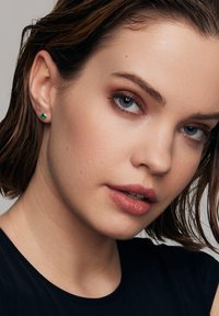 Szmaragdowe kolczyki wkrętki osadzone w srebrze, charakteryzujące się kwadratowym szlifem, prezentowane na neutralnym tle, dopełnione gładką teksturą skóry.