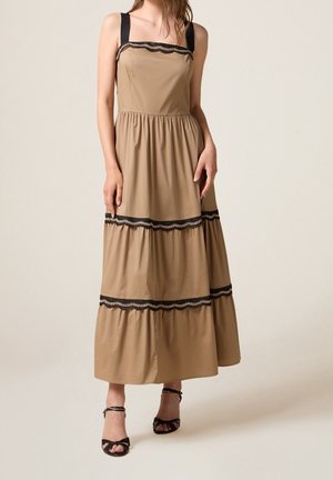 Vrouw die een mouwloze beige maxi-jurk draagt met zwarte kanten afwerking en zwarte enkellaarsjes met bandje tegen een effen achtergrond.