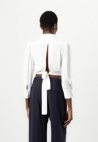 Blusa bianca corta con maniche lunghe, apertura a occhiello nella parte posteriore e dettaglio della vita legato, abbinata a pantaloni navy a vita alta. Tessuto dalla superficie liscia.