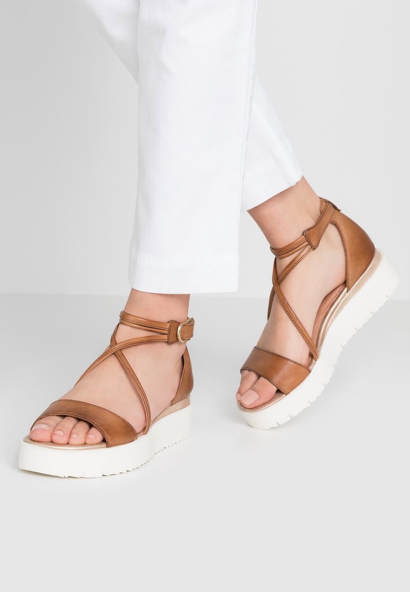 Sandales en cuir marron avec un design de sangles entrecroisées, boucle de cheville et semelle en plateforme blanche, mettant en valeur une texture lisse et un bout ouvert.
