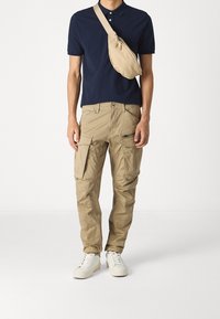 Marinblå polo skjorta, beige cargo byxor med flera fickor, vita sneakers. Väskan är beige, bärs över kroppen, med dragkedjestängning.