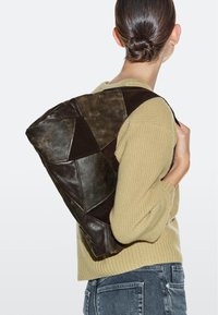 Person mit einem Dutt, die einen beigen Pullover und blaue Jeans trägt und eine große dunkelbraune Schultertasche aus Leder mit Patchwork-Design bei sich trägt.