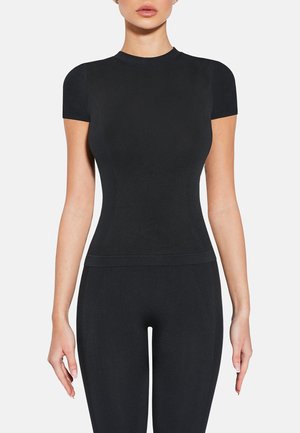 Kobieta ubrana w dopasowany czarny top z krótkim rękawem i pasujące czarne legginsy, stojąca na tle jednolitego białego tła.