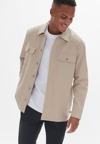 Chaqueta beige de botón con cuello, dos bolsillos en el pecho y botones marrones, llevada sobre una camiseta blanca. Tela suave y ajuste relajado.